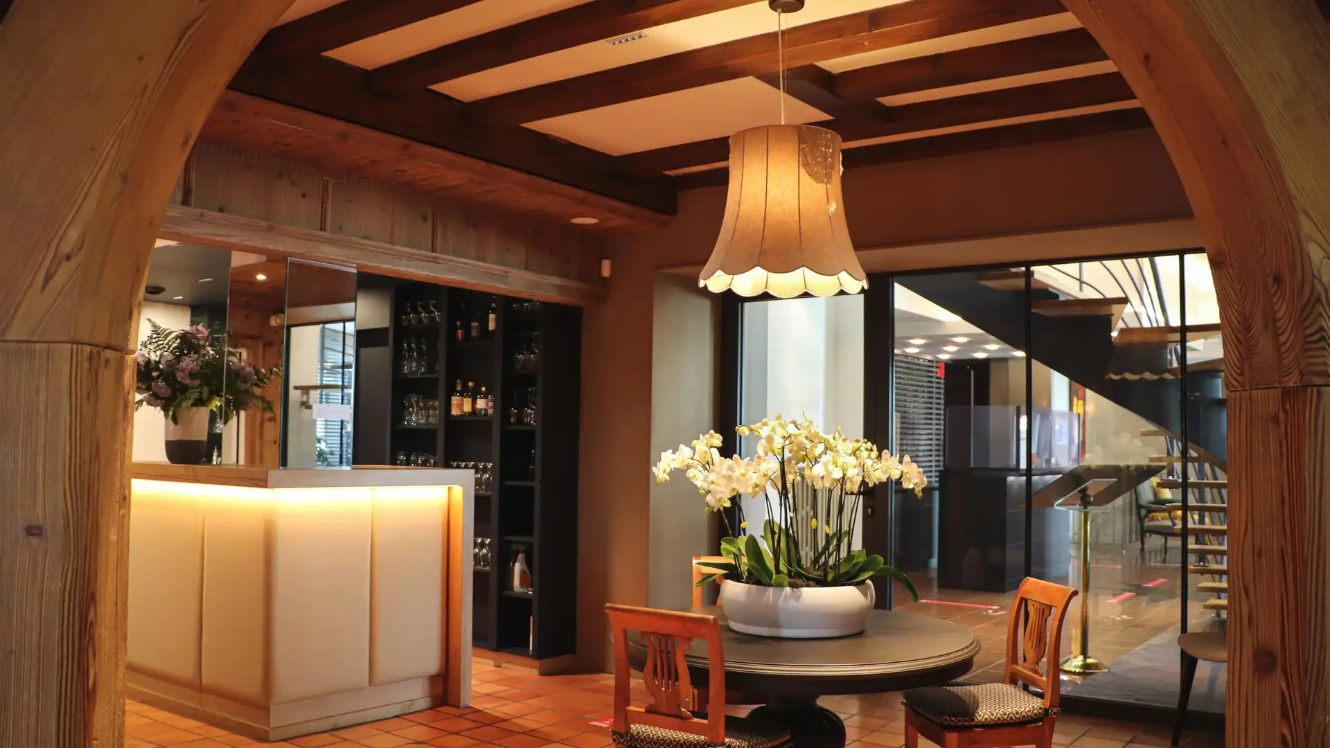 A restaurant interior with a reception counter, tables, chairs, and decorative plants under a wooden ceiling with a hanging lamp.