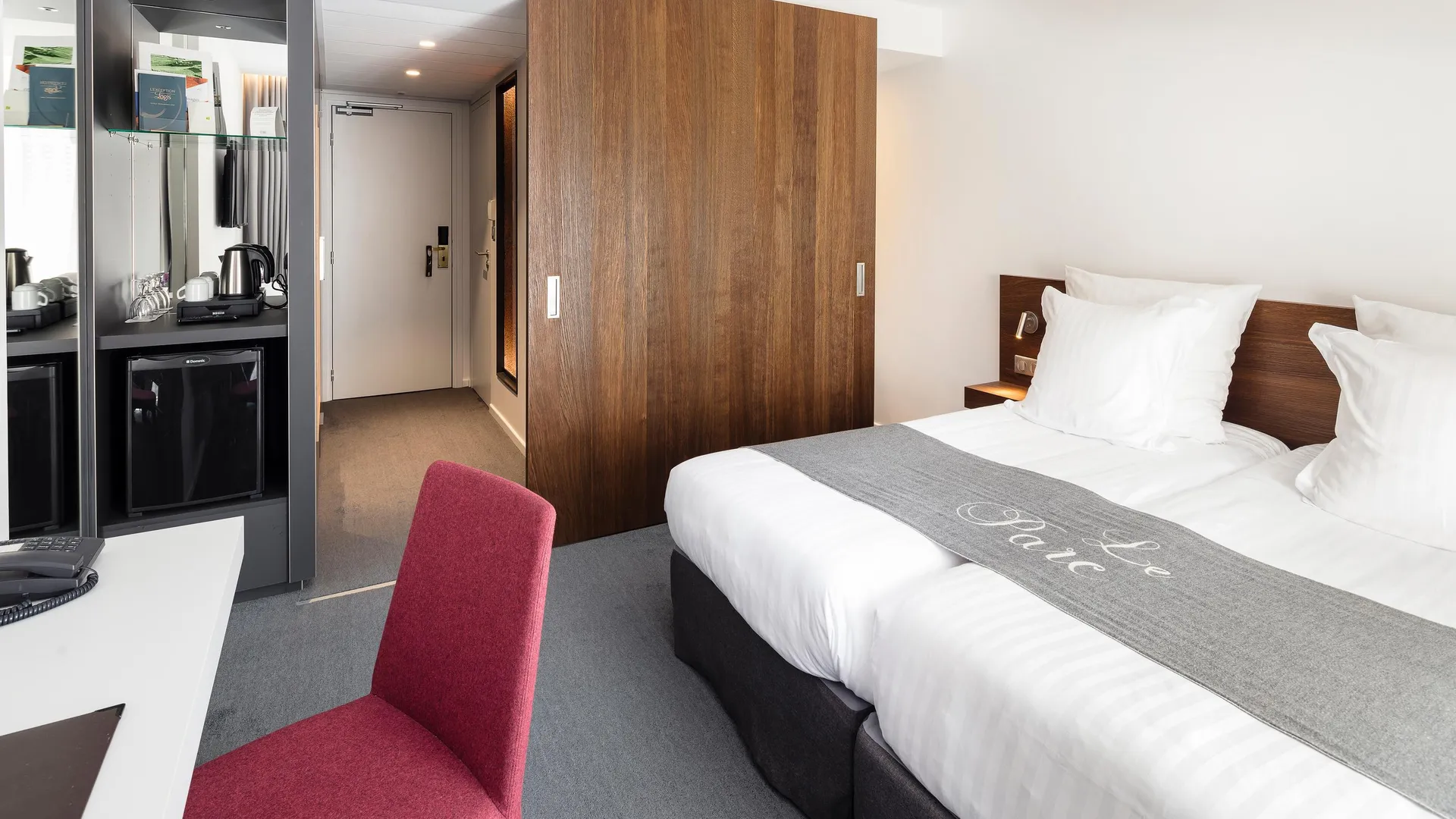 Hotel room with a double bed, a red chair, a mirror, and a visible mini-fridge in the corner.