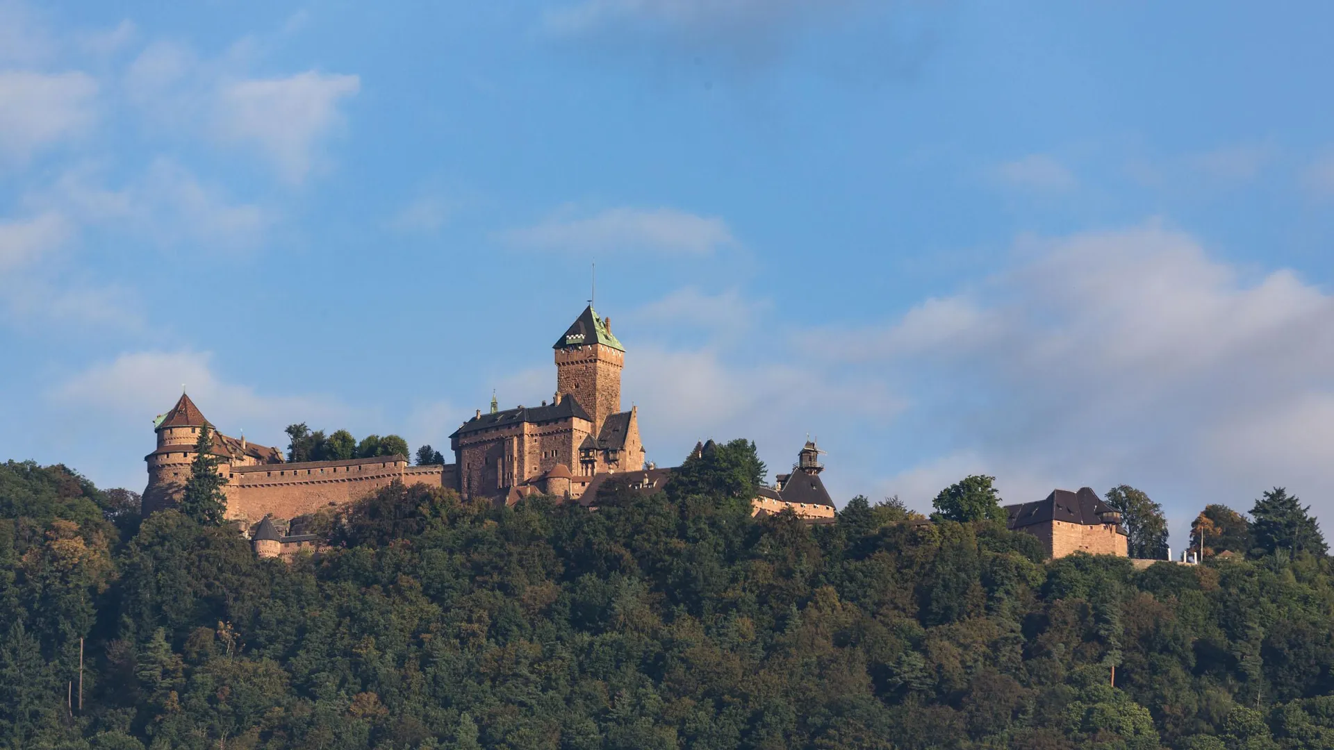 Château médiéval perché sur une colline boisée sous un ciel partiellement nuageux