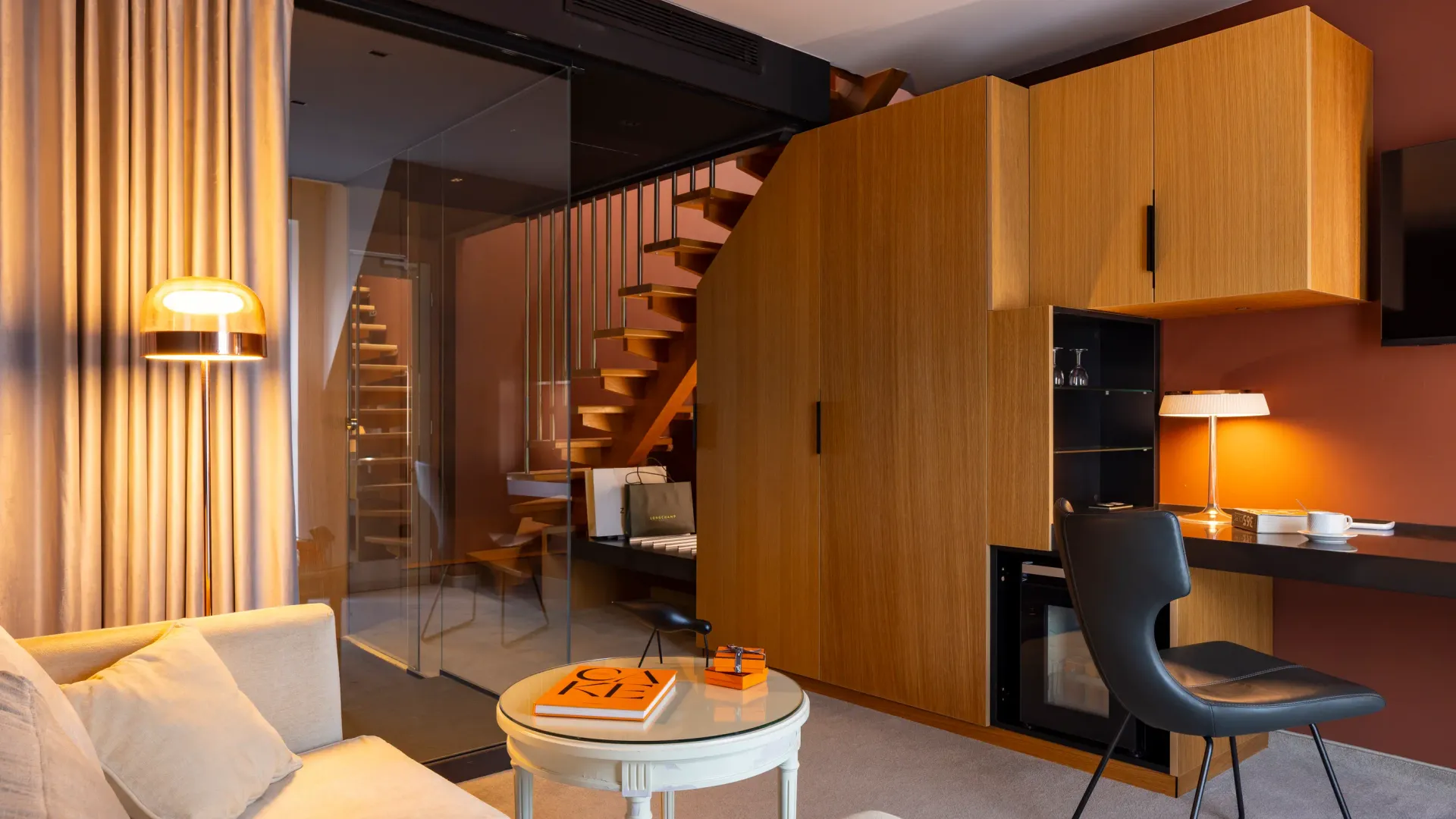 Salon moderne avec canapé beige, table basse, bureau et escalier en verre menant à l'étage