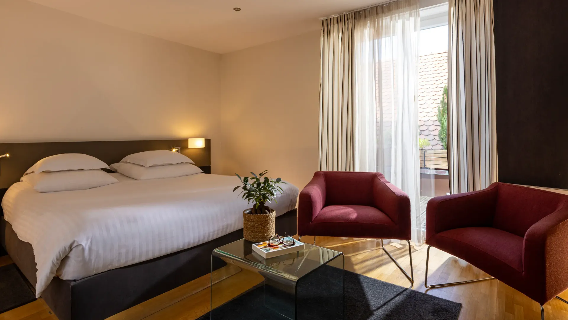 Zimmer mit einem großen Doppelbett, zwei bordeauxroten Sesseln, einem Glastisch und einem großen Fenster mit Vorhängen